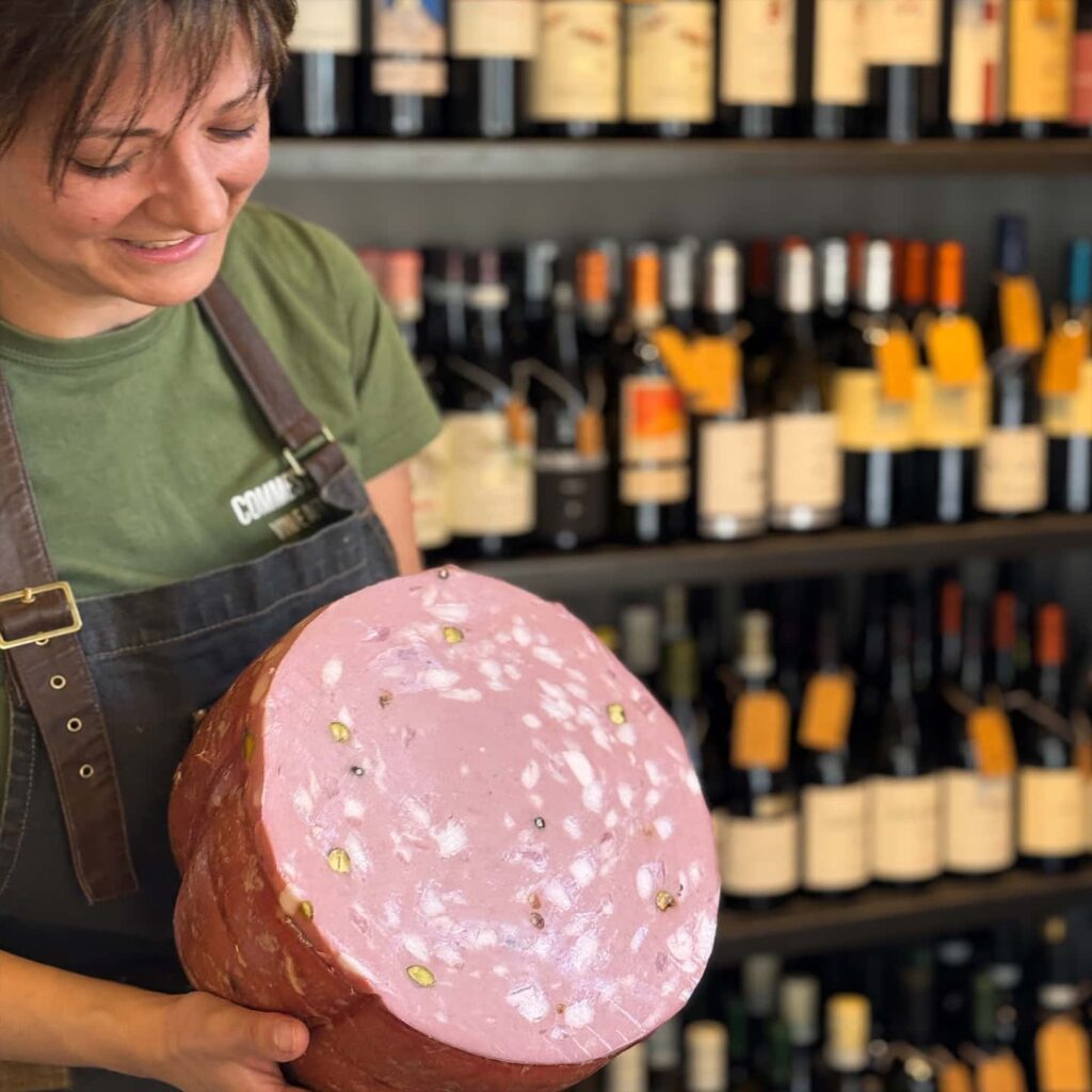 Commessa taglia una mortadella. Sullo sfondo delle bottiglie di vino